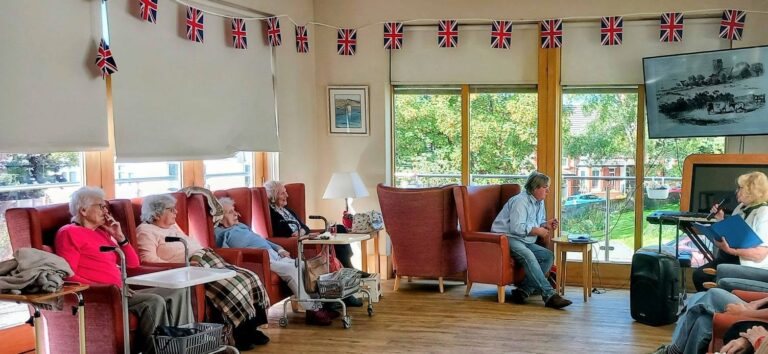 A group of elderly residents sat in armchairs in a lounge decorated with Union Jack bunting, enjoying a Saturday afternoon listening to a woman reading aloud.