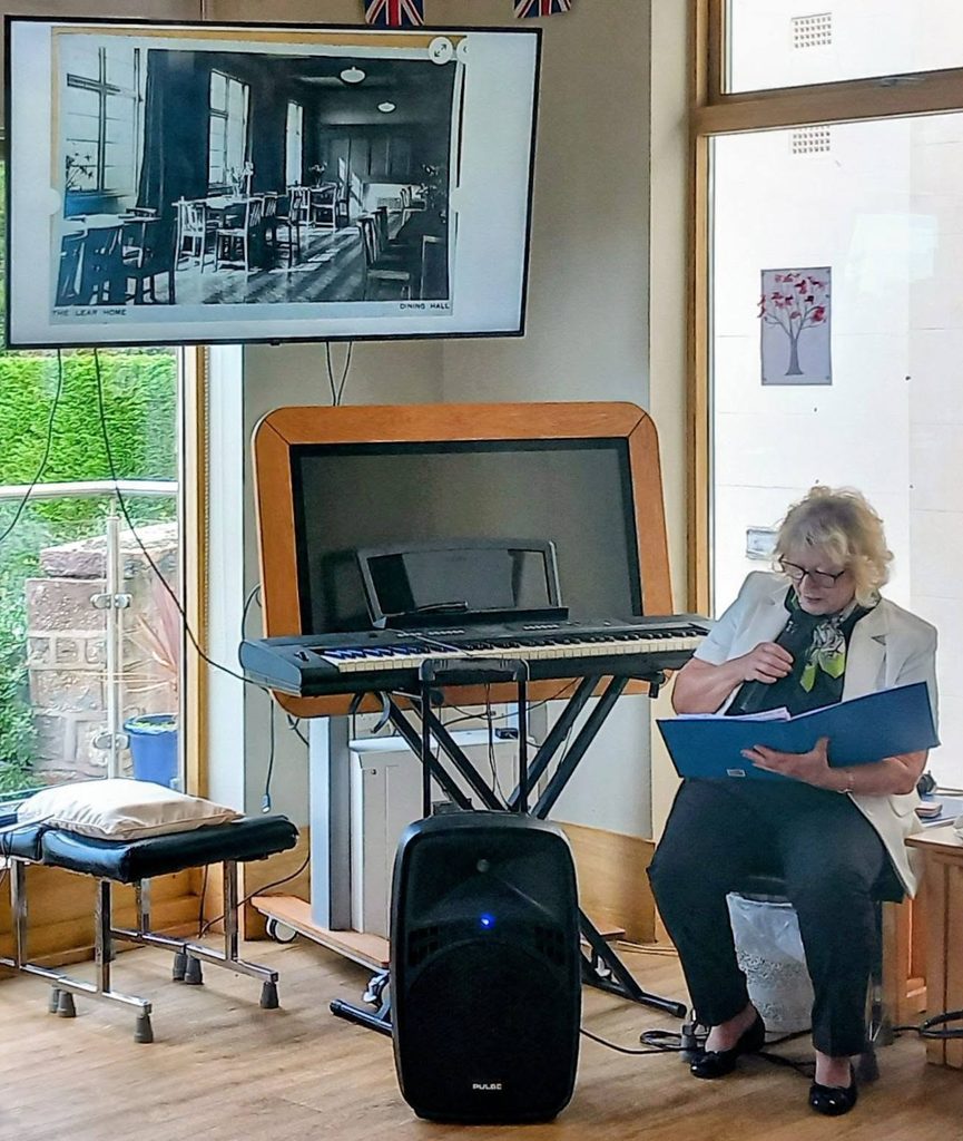 A blonde woman sits in a chair, reading from a blue folder into a microphone before a keyboard and a screen displaying a Saturday time-travel photograph of a vintage dining hall.
