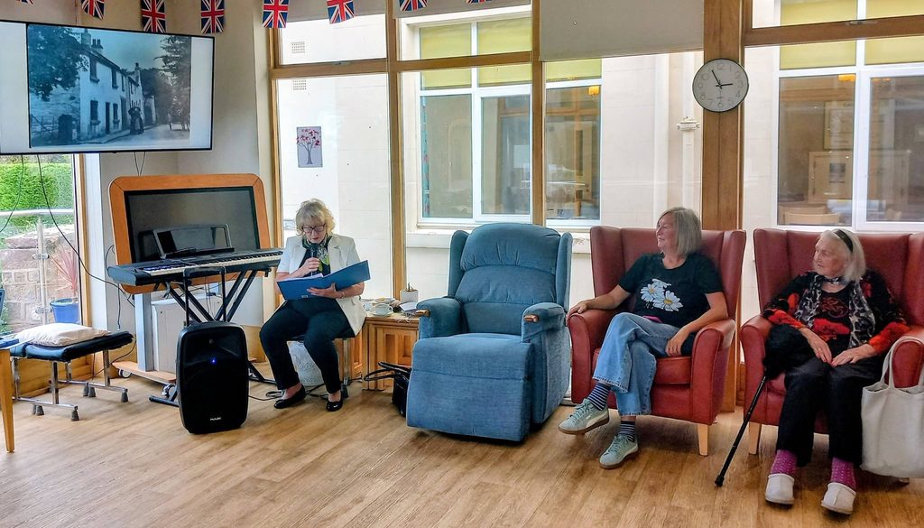 A woman speaks into a microphone whilst two others sit in armchairs in a community room decorated with Union Jack bunting for a trip through time.