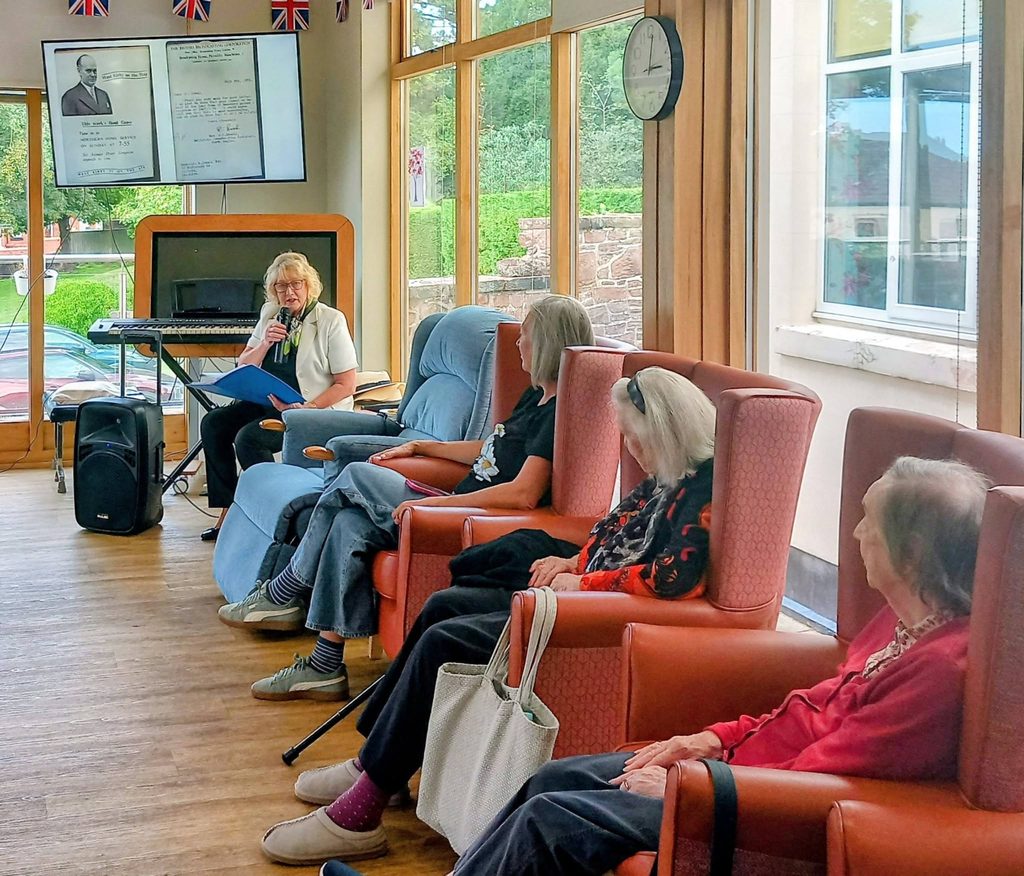 A woman stands at a microphone speaking to a group of seated residents in a lounge during a Saturday afternoon trip through time.