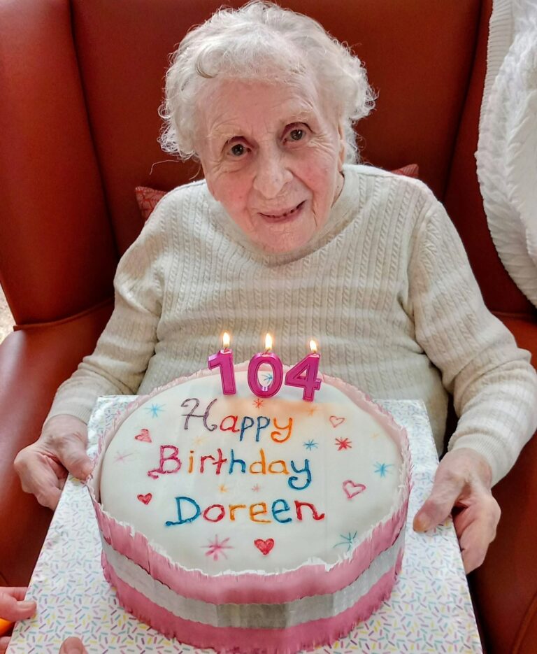 An elderly woman holds a birthday cake featuring candles that spell 104 and an inscription that reads Happy Birthday Doreen.