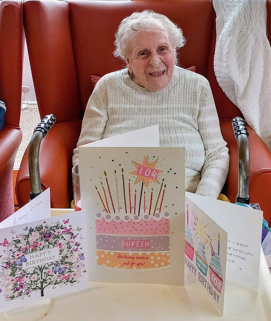 Doreen smiling in an armchair with three birthday cards displayed on a table in front of her; the central card features a cake and the number 104.