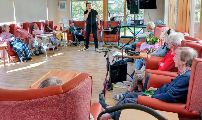 Jay Irving sings into a microphone for elderly residents seated in armchairs in the Lear House communal lounge.