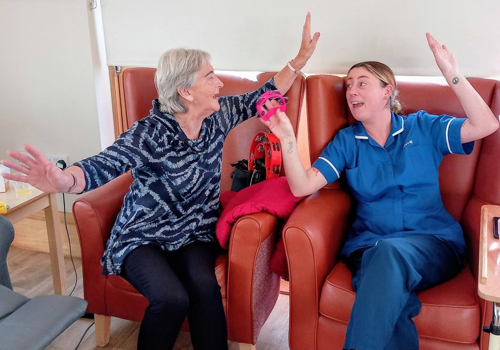 An elderly woman and a care worker in a blue uniform sitting in armchairs, high-fiving whilst holding a small pink tambourine during a Lear House music event.