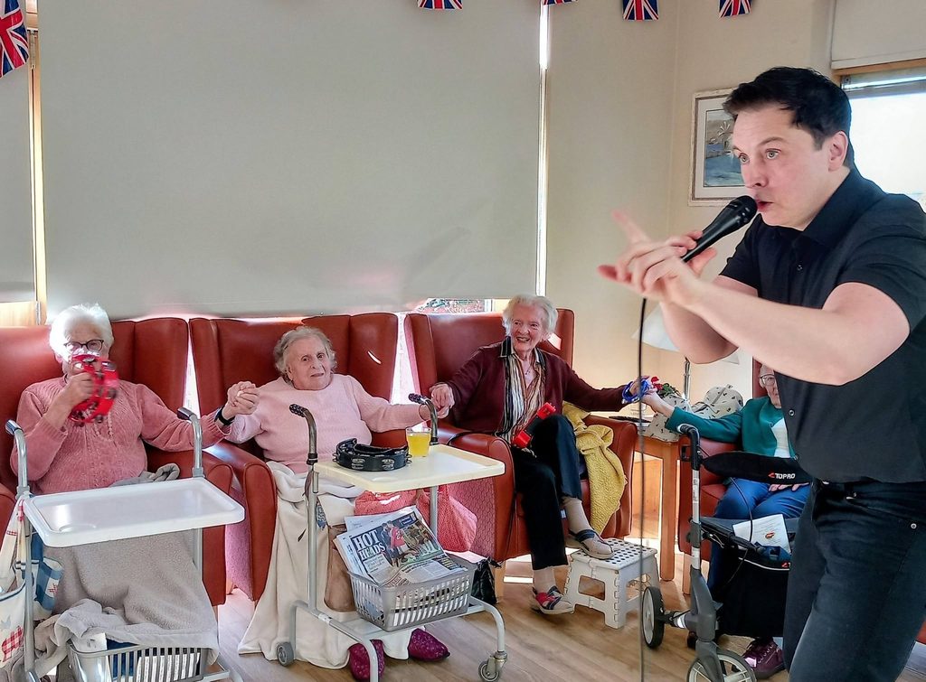 A man in a black shirt singing into a microphone for a group of elderly residents who are dancing in their armchairs whilst holding hands and playing percussion instruments.
