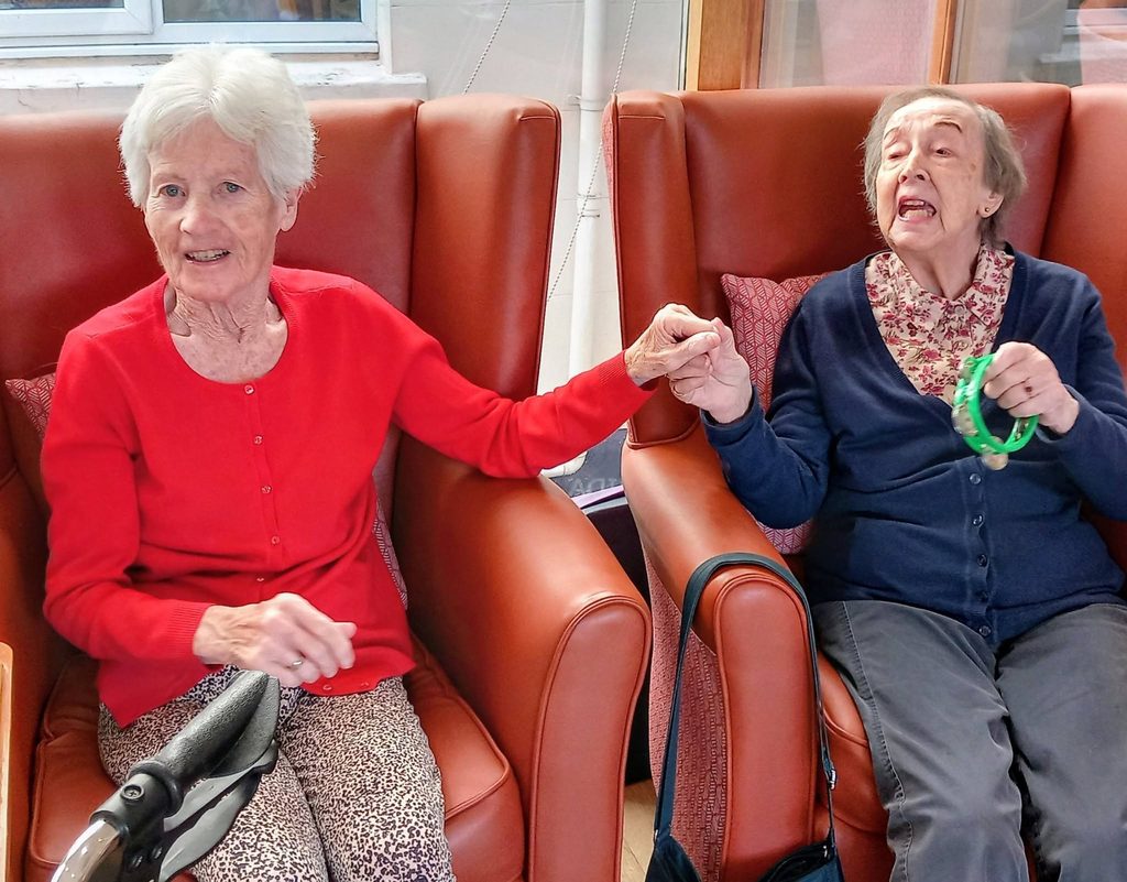 Two elderly women sit in armchairs at Lear House, holding hands during a music performance.