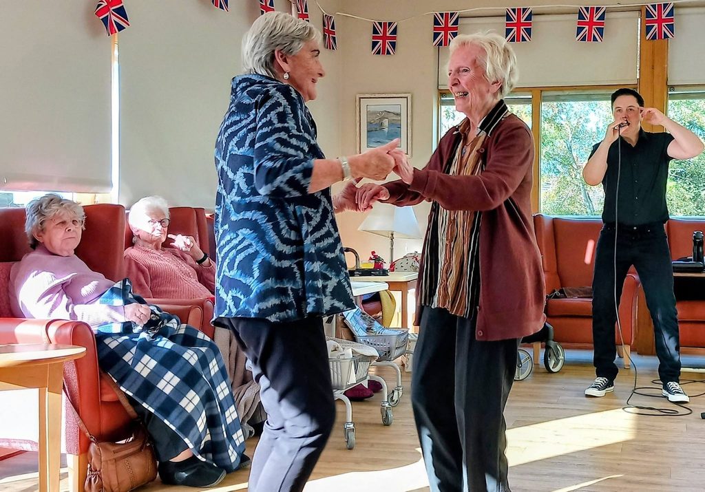 Two older women dance in a room decorated with bunting whilst Jay Irving performs live music on the microphone nearby.