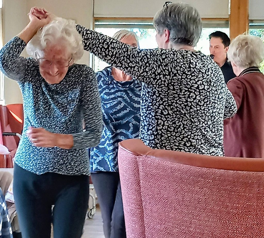 Older women dance to music in a room whilst a man stands in the background at the Lear House.