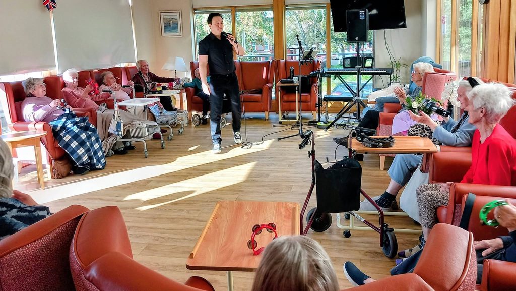 Jay Irving performs with a microphone for residents seated in armchairs at Lear House, some using mobility aids.