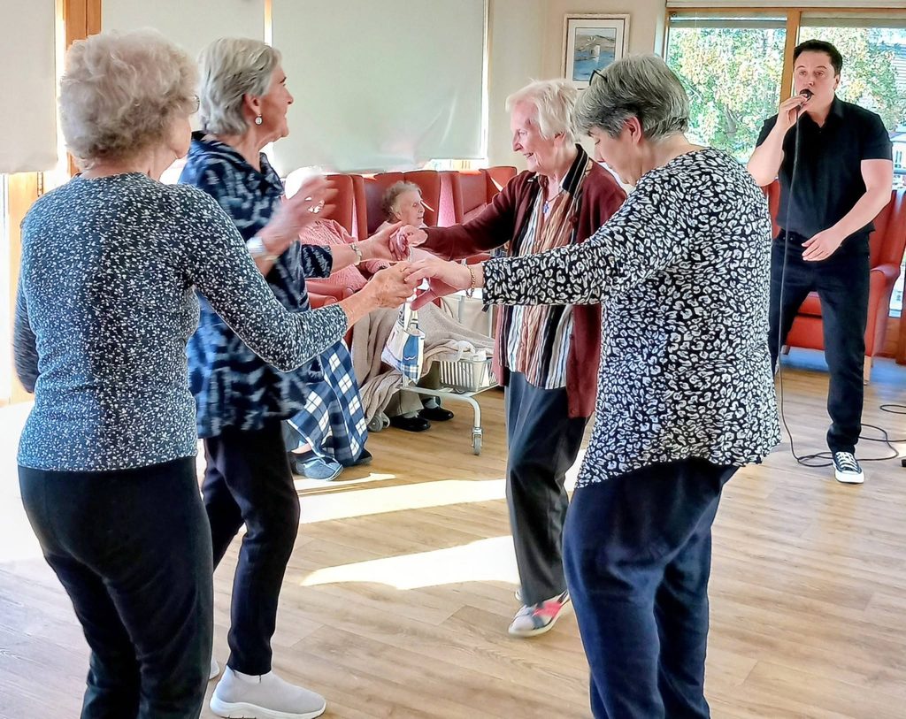 Four older women hold hands and dance in a circle whilst Jay Irving sings into a microphone during a live music performance at Lear House.