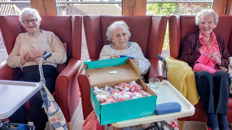 Three elderly women from a Knit and Natter craft group sitting in armchairs, one holding a shoebox filled with small, wrapped presents.
