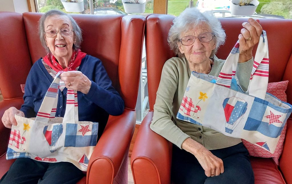 Two elderly women sitting in leather armchairs during a knit and natter, each holding up a fabric tote bag decorated with patterns of hot air balloons and picnic items.