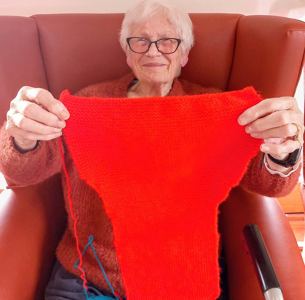An elderly woman with white hair and glasses sits in a chair at a craft group, holding up a piece of bright red knitting.