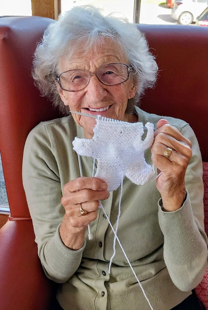 An elderly woman sits in a chair, smiling whilst holding knitting needles and a piece of white fabric during a knit and natter session.