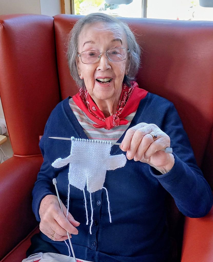 An elderly woman sits in a red armchair, smiling whilst knitting a white garment during a Knit and Natter group.