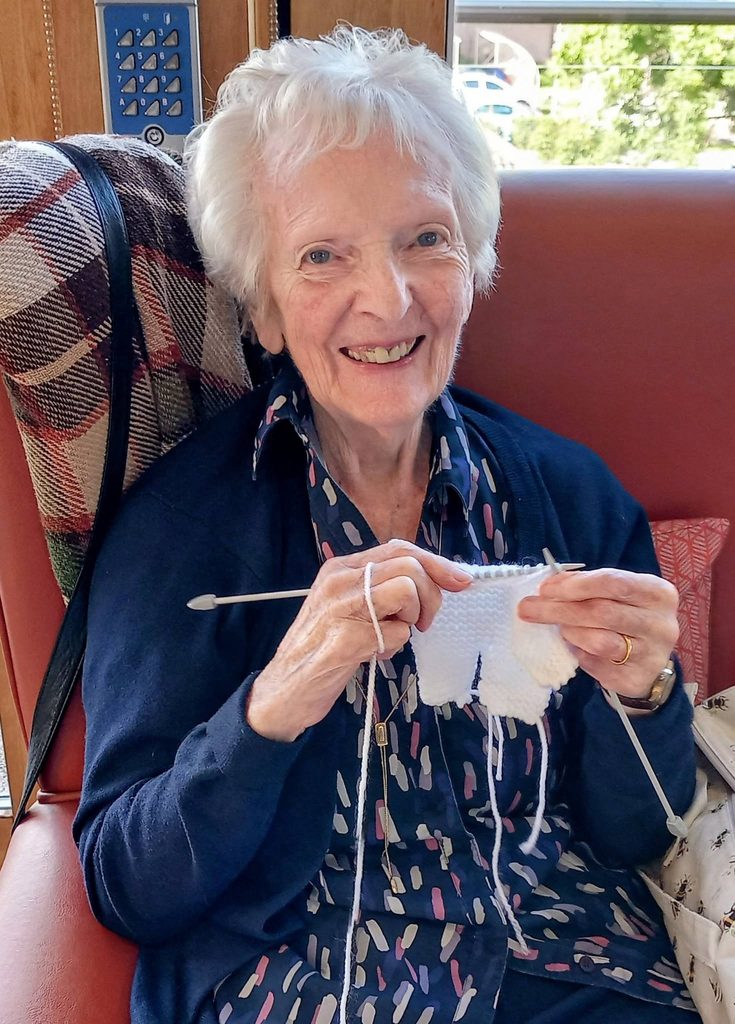An elderly woman smiling whilst knitting with white wool at a Knit and Natter craft group.
