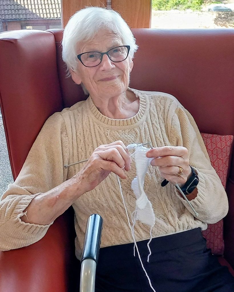 An elderly woman with white hair and glasses sits in a red armchair, knitting with white wool as part of a knit and natter group.
