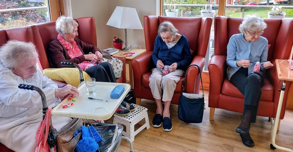 Four elderly women sit in red armchairs in a craft group, each working on a small project with their hands.