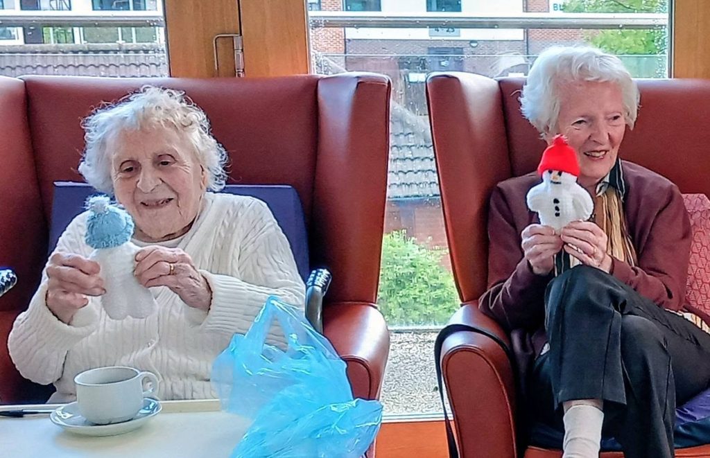 Two elderly women sitting in armchairs, each holding a handmade snowman during a knit and natter session.