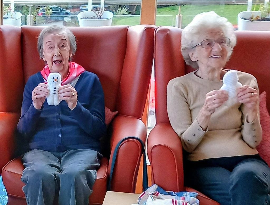 Two elderly women sit in red armchairs at a Knit and Natter, each holding a small white knitted snowman.