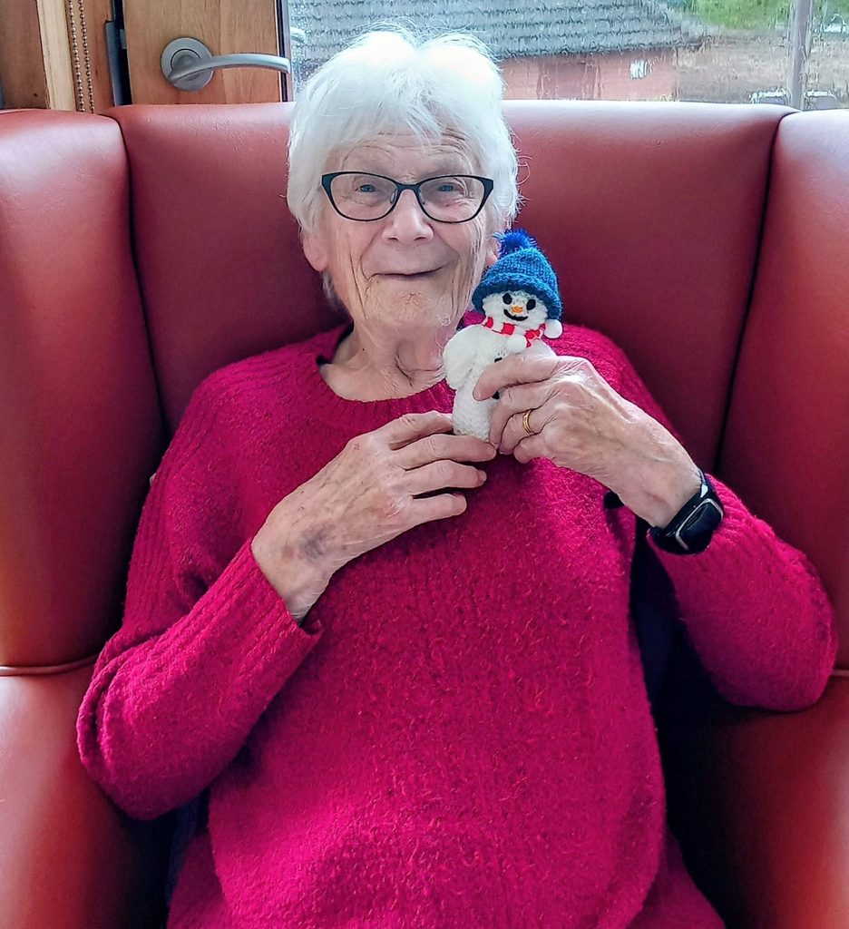 An elderly woman with white hair and glasses wearing a red jumper, sitting in a red armchair and holding a small knitted snowman during a Knit and Natter session.