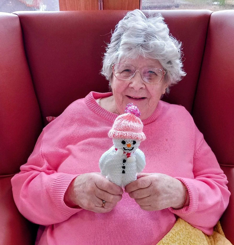 An elderly woman wearing a pink jumper sits in a red armchair, holding a handmade snowman during a knit and natter session.