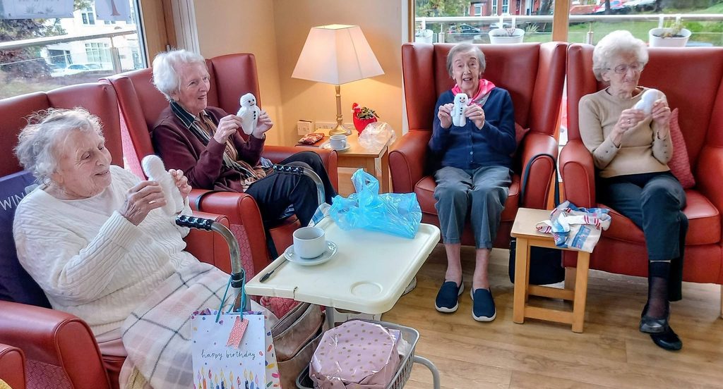 Four elderly women sit in leather armchairs during a Knit and Natter session, each holding a small plush snowman.