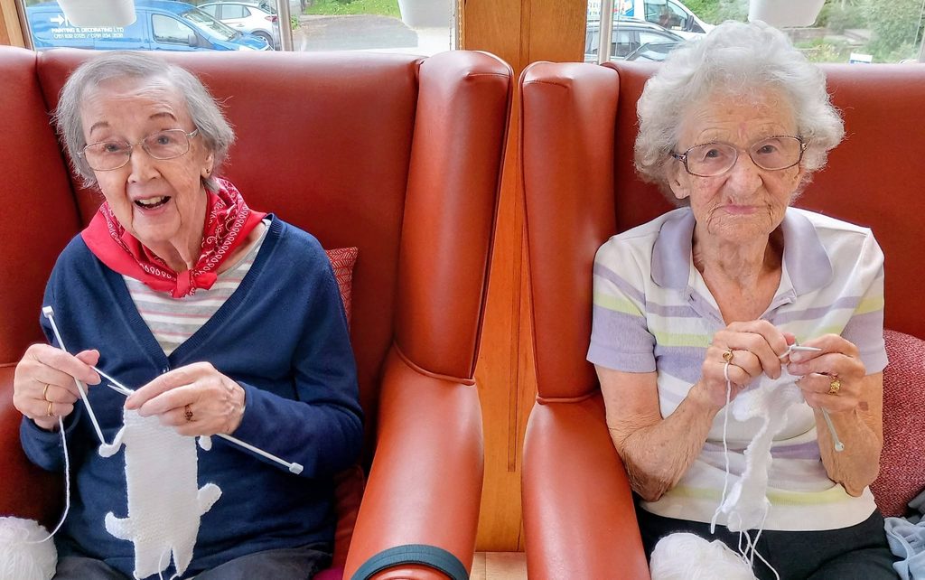 Two elderly women sitting in leather armchairs at the Christmas Tree Festival, both knitting with white wool.