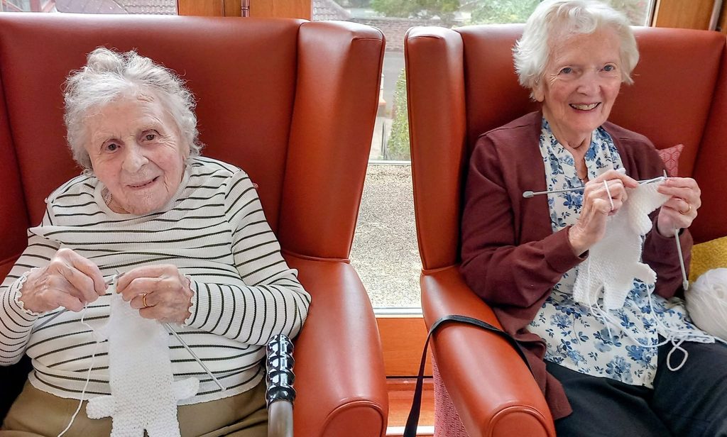 Two elderly women sitting in leather armchairs at a Knit and Natter, both knitting white items.