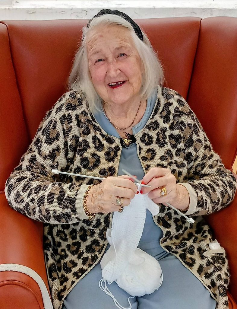 An elderly woman wearing a leopard-print cardigan sits in a red armchair, knitting with white wool during a Knit and Natter session.