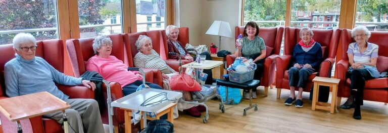 Seven people sit in red armchairs in a sunlit room for a Knit and Natter, with one woman holding up a small present.