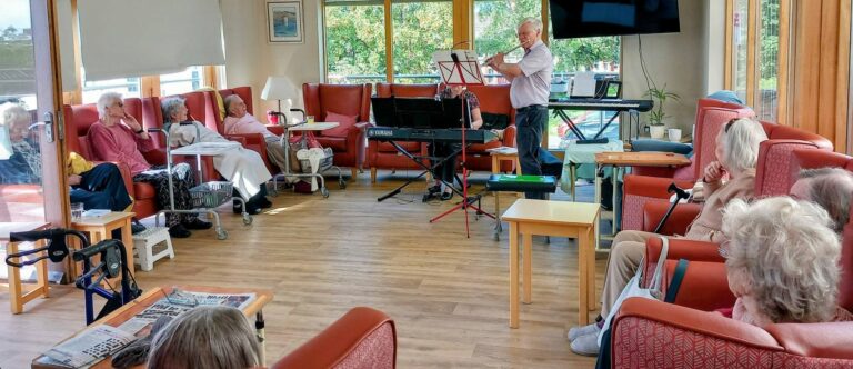 A man performs a live flute recital for a group of elderly residents seated in armchairs in a communal lounge.