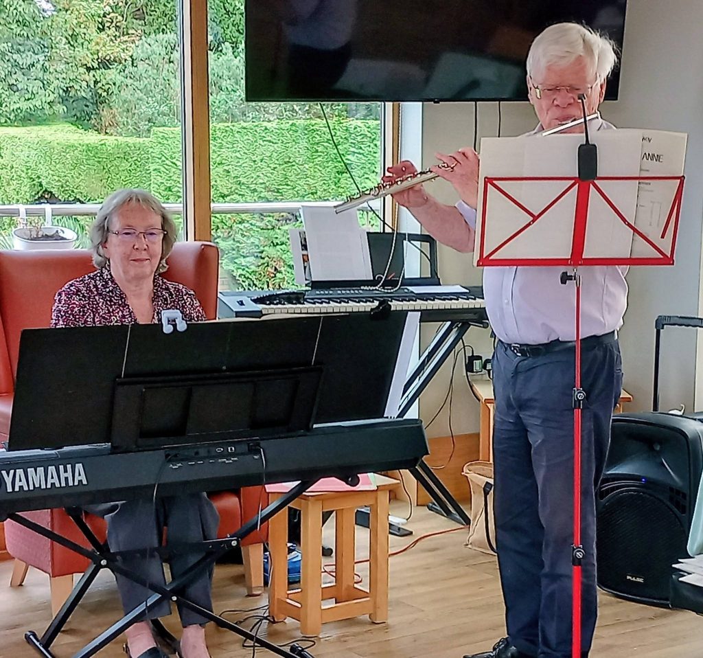A woman plays the piano whilst a man stands playing the flute, both reading from sheet music on stands during a concert.