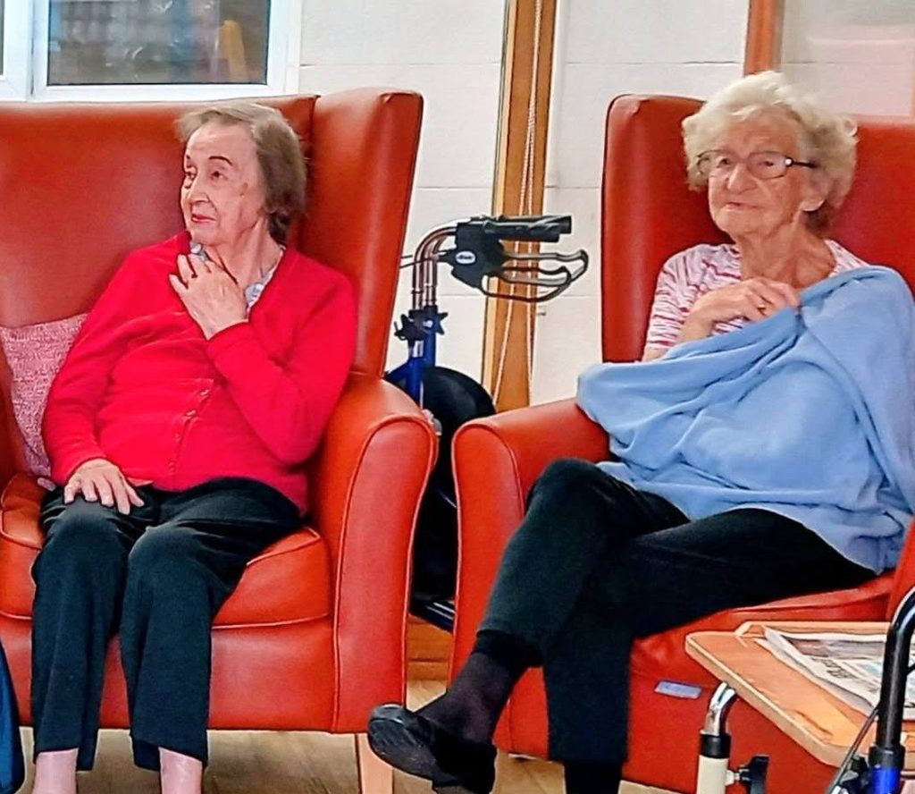 Two elderly women sitting in orange armchairs during a piano and flute performance.