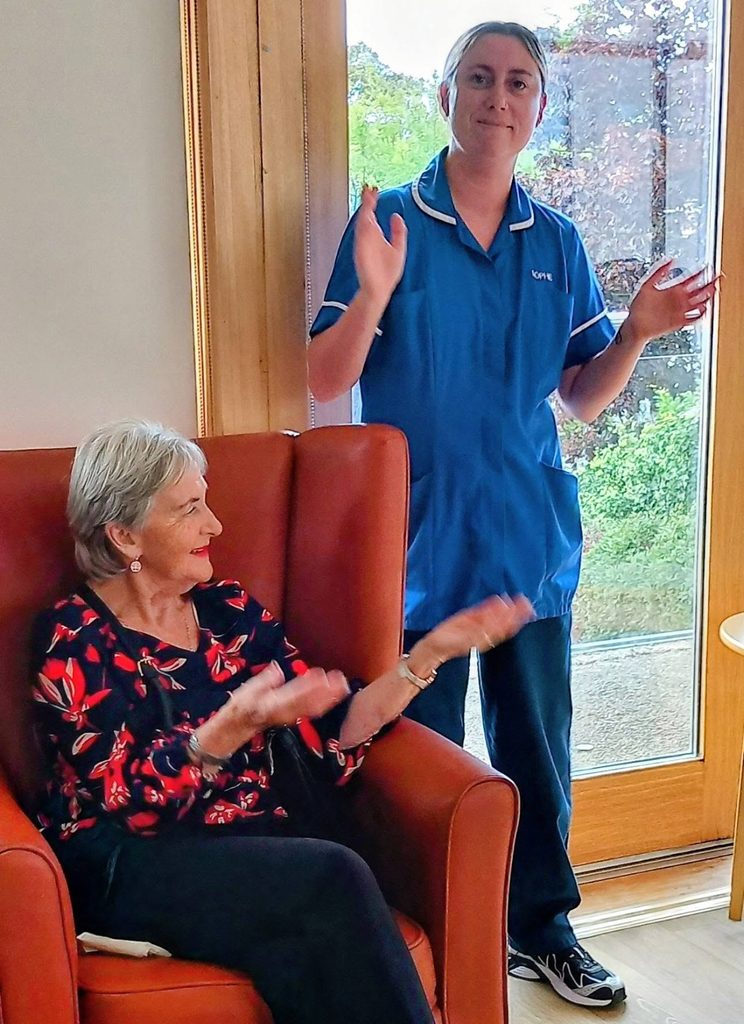An elderly woman sitting in an armchair and a carer in a blue uniform both clapping their hands after a musical performance.