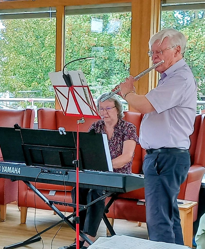 A man playing the flute and a woman playing an electronic keyboard during a concert in front of large windows.