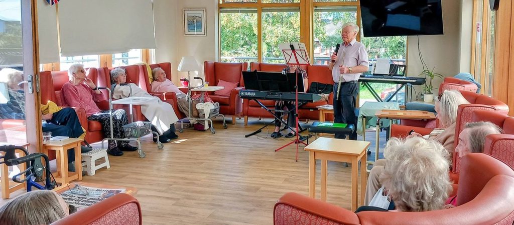 A man plays the flute for an audience of elderly people sitting in a circle of armchairs in a communal room during a concert.