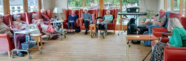 A group of elderly people sit in armchairs in a circle listening to retired police officer Chris Pierce speak into a microphone.