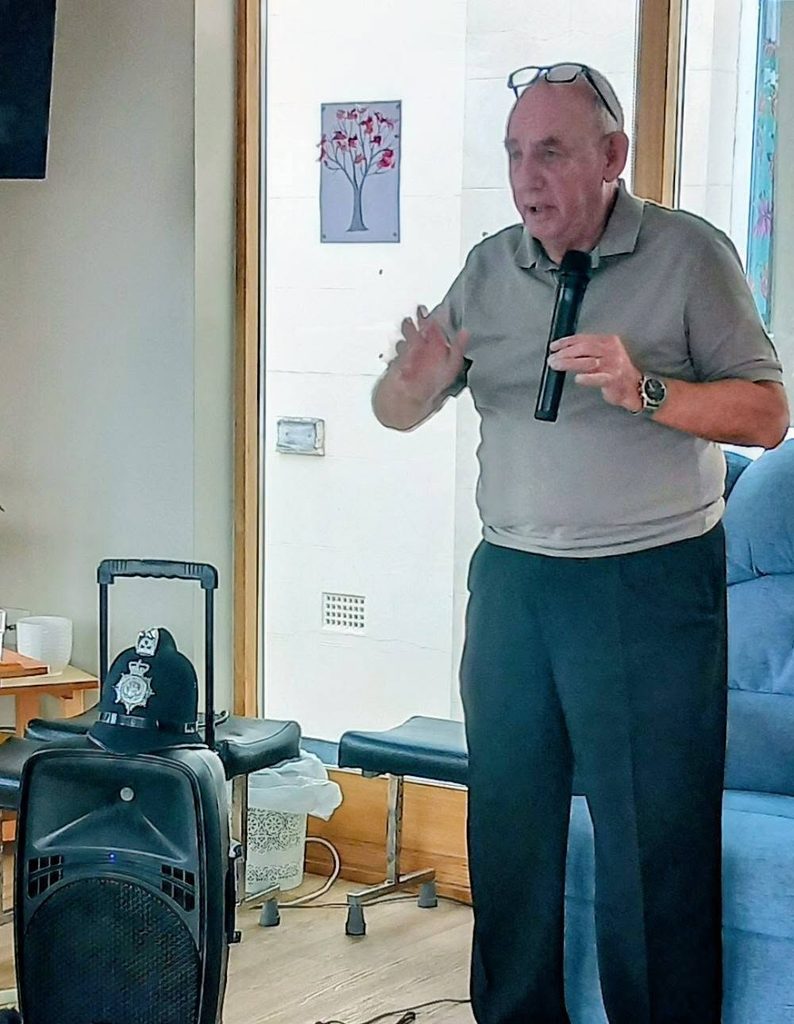 Retired police officer Chris Pierce stands holding a microphone, with a police helmet resting on a loudspeaker in the foreground.