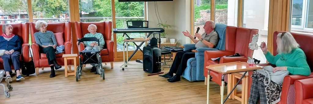 A group of older adults sit in armchairs in a lounge whilst retired police officer Chris Pierce speaks into a microphone.