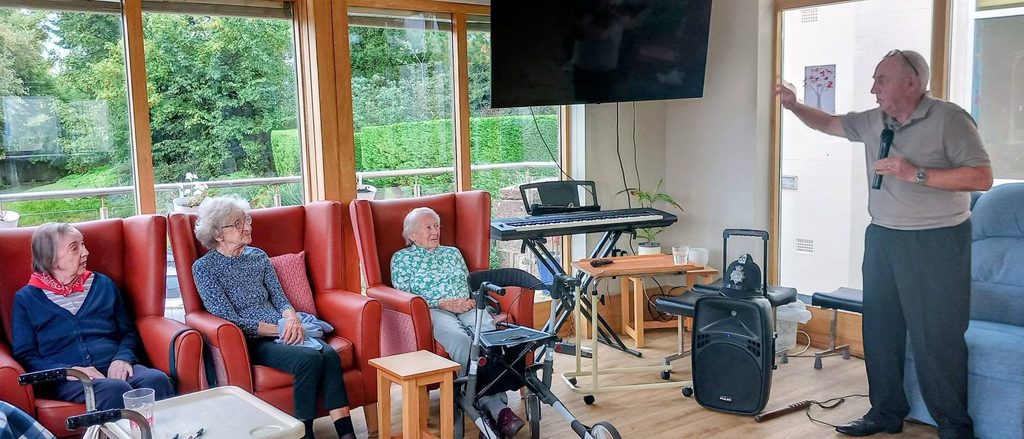 Retired police officer Chris Pierce holds a microphone and gestures towards a wall-mounted television whilst three elderly residents sit in armchairs nearby.