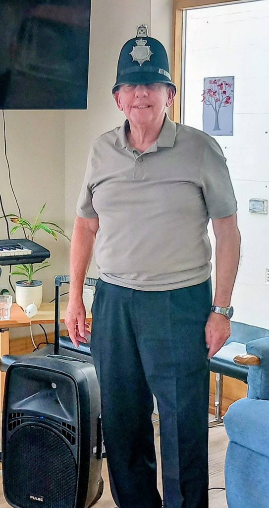 Retired police officer Chris Pierce smiles whilst wearing a traditional British police helmet.