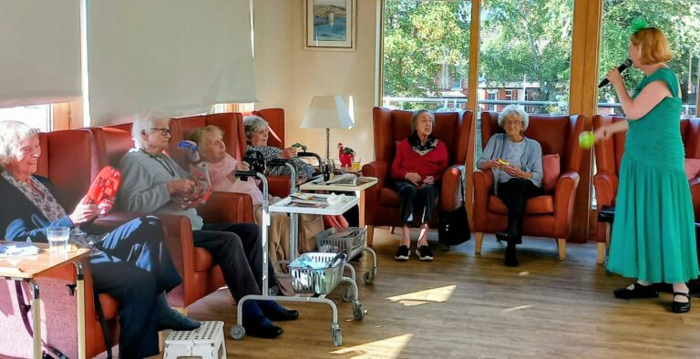 A woman in a green dress sings into a microphone for a group of elderly residents seated in armchairs, some of whom are playing tambourines.