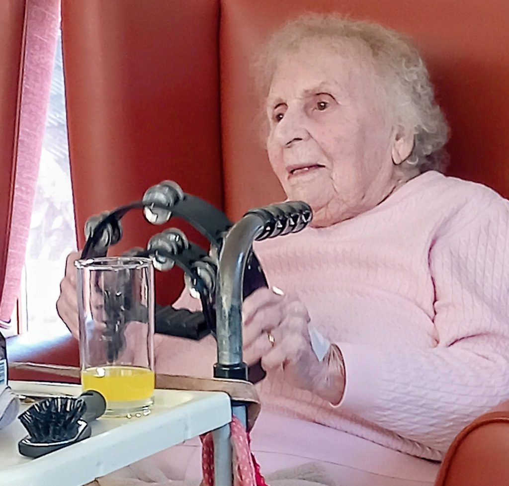 An elderly woman sits in a red armchair, singing whilst holding a tambourine.
