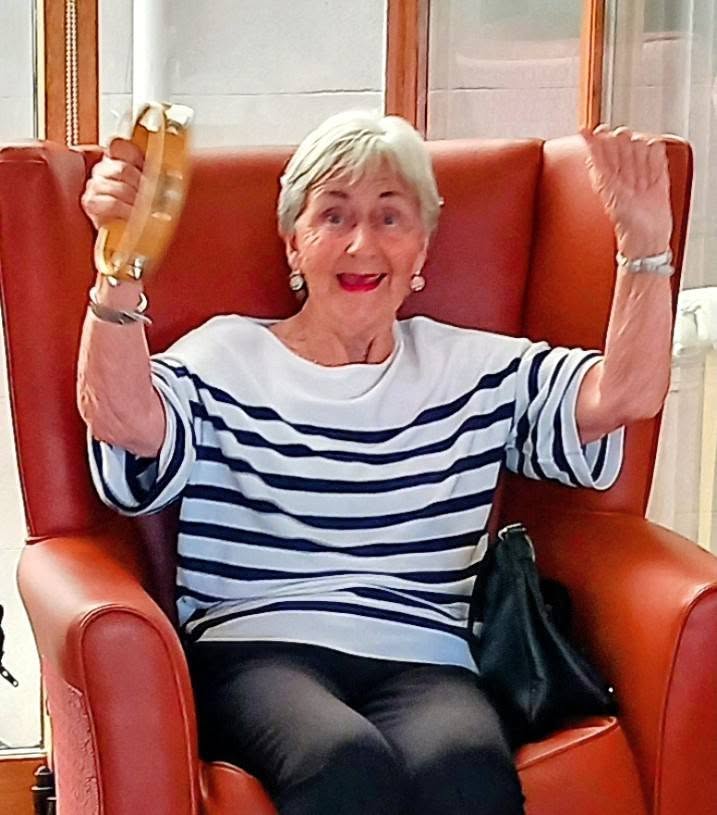 An elderly woman sits in a high-backed armchair, smiling and singing whilst holding a tambourine aloft with one hand.
