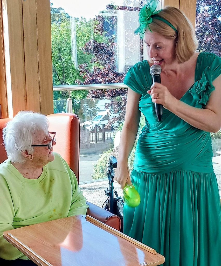 A woman in a green dress sings into a microphone whilst dancing and holding a maraca for an elderly woman seated in a chair.