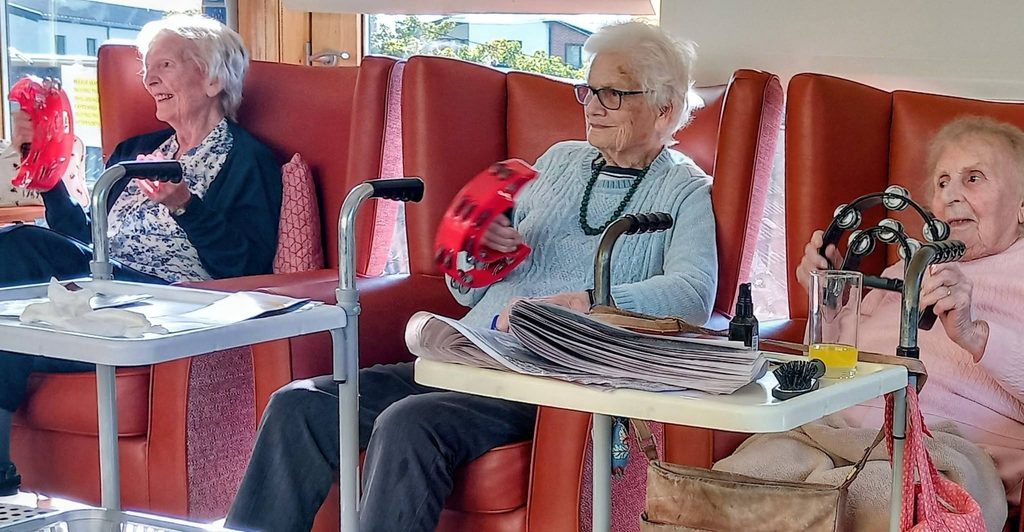 Three elderly women sit in armchairs, singing whilst each plays a tambourine.