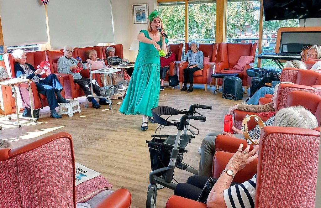 A performer in a green dress sings into a microphone in a lounge while elderly residents seated in armchairs play tambourines.