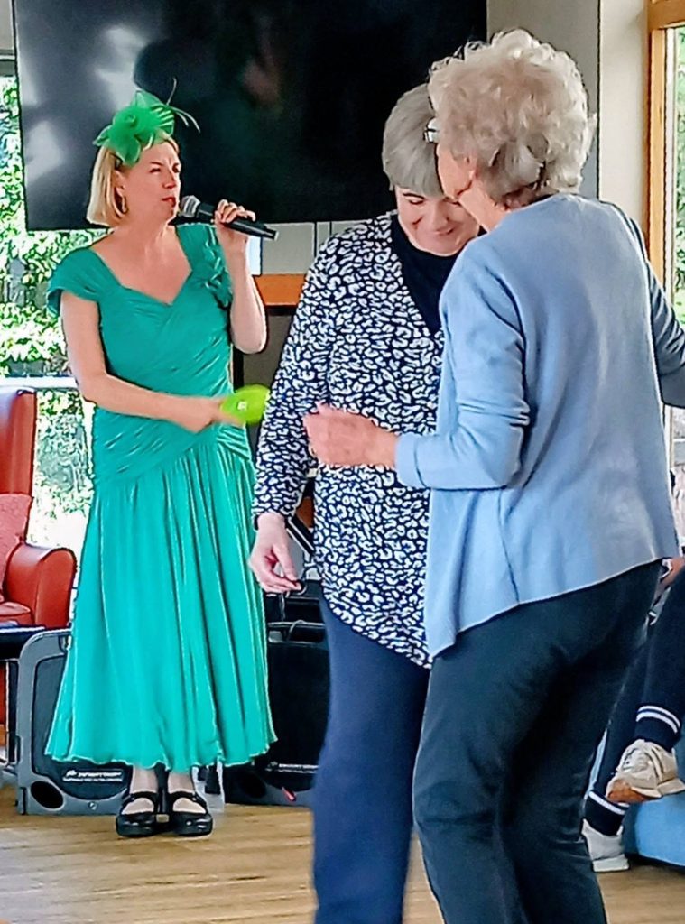 A woman in a green dress and matching fascinator sings into a microphone whilst two women dancing in front of her play tambourines.