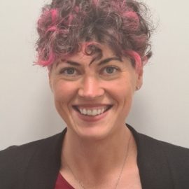 A person with short, curly hair featuring pink highlights, dressed in a burgundy top, black blazer, and a necklace, smiling at the camera.